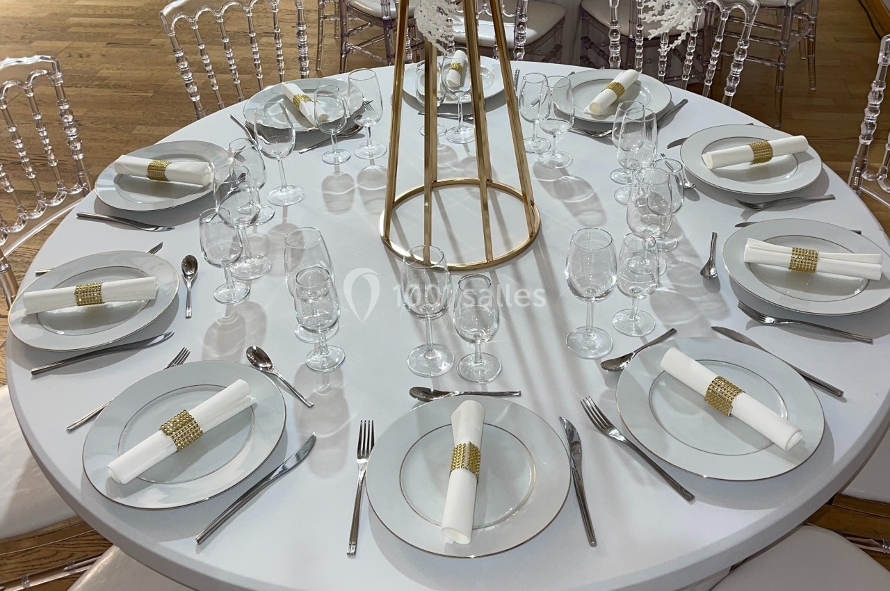 Table ronde élégamment dressée avec nappes blanches, assiettes, couverts, verres et serviettes ornées d'anneaux dorés.