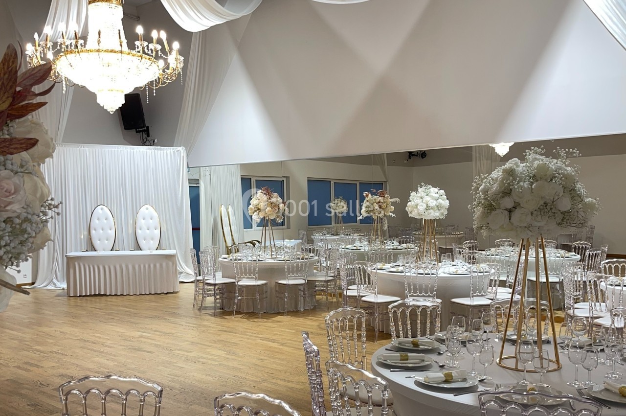 Salle de réception élégante avec tables décorées de fleurs blanches et chaises transparentes sous un grand lustre.