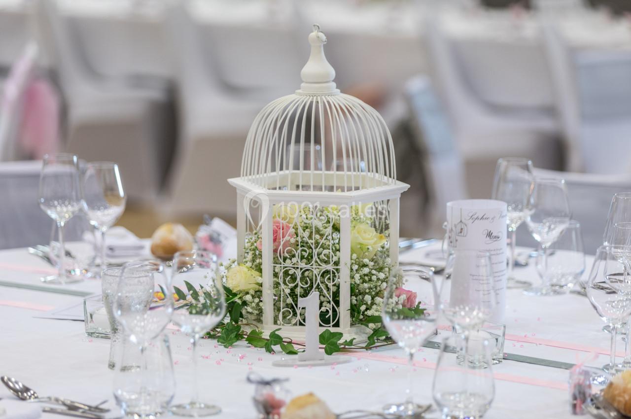 Centre de table avec une cage blanche décorée de fleurs roses et blanches, entourée de verres et couverts.
