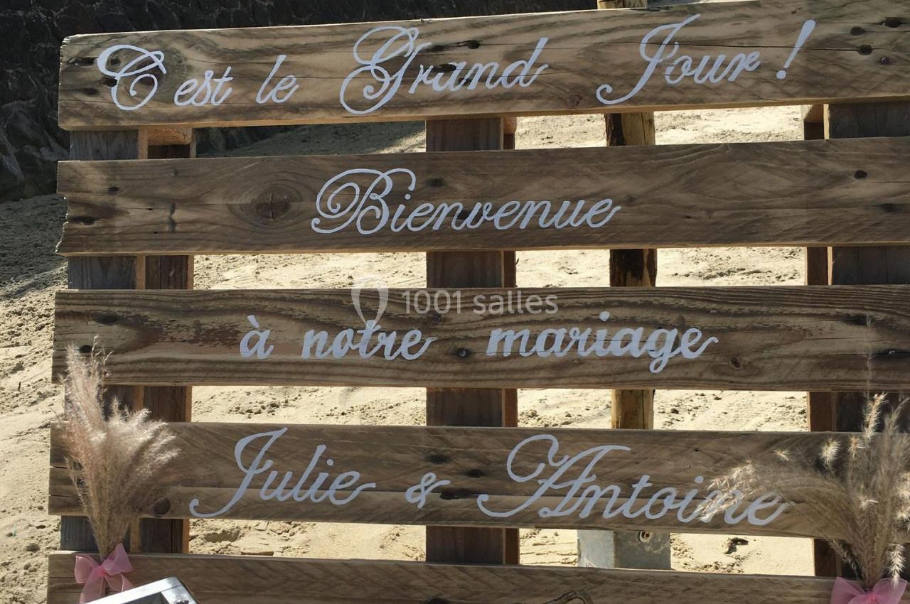 Panneau en bois sur une plage, décoré de textes peints annonçant un mariage et les prénoms Julie et Antoine.