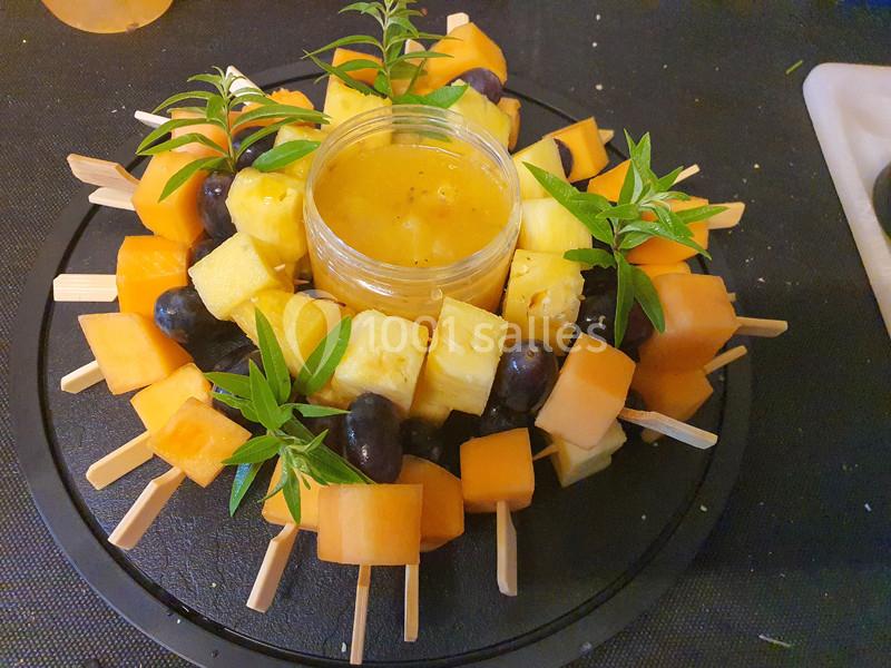 Brochettes de fruits variés autour d'un pot de sauce, décorées de feuilles vertes sur un plateau noir.