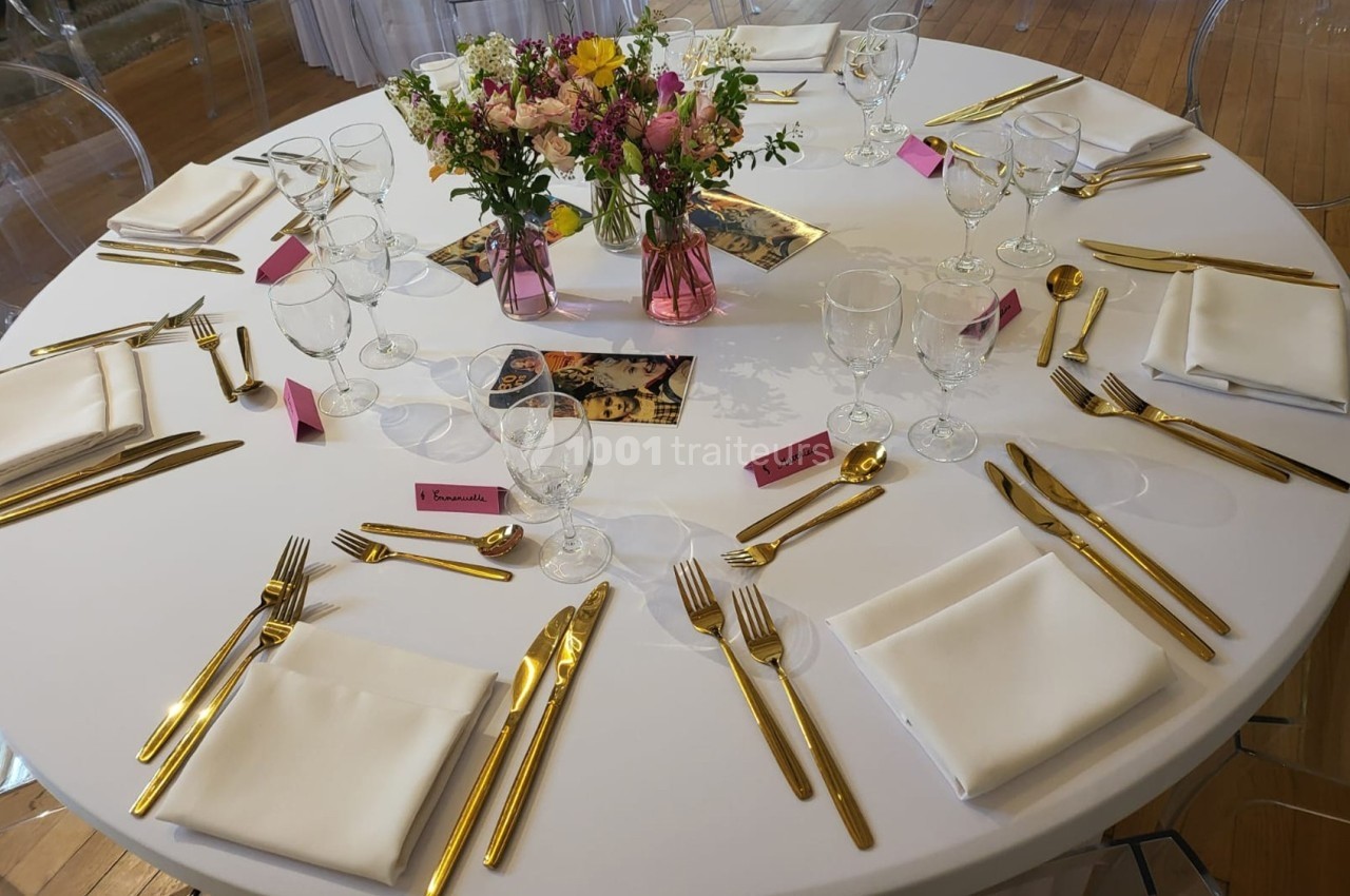 Table ronde dressée avec nappes blanches, couverts dorés, serviettes pliées, verres et centre de table floral coloré.