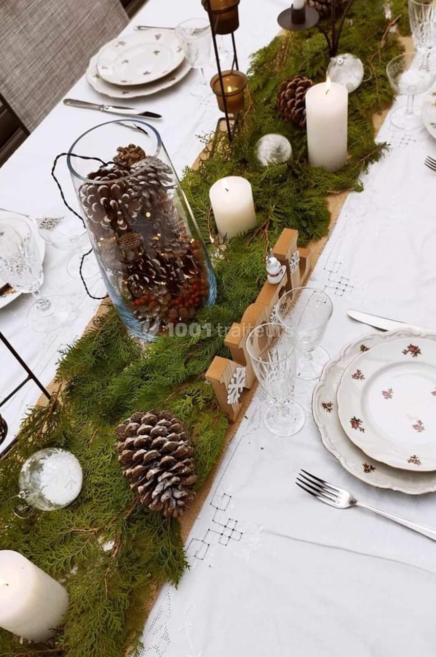 Table décorée pour un repas, ornée de bougies, pommes de pin, branches de sapin et vaisselle élégante.