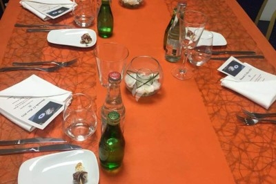 Table dressée avec nappe orange, couverts, verres, bouteilles et assiettes, disposée pour un repas ou une réunion.