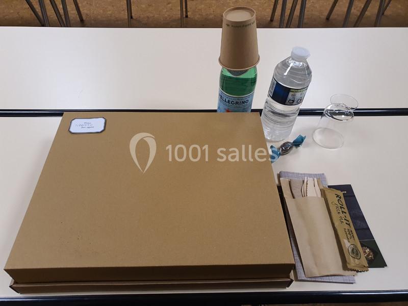 Repas emballé avec boîte en carton, bouteille d'eau, gobelets empilés, couverts en bois et serviette sur une table blanche.