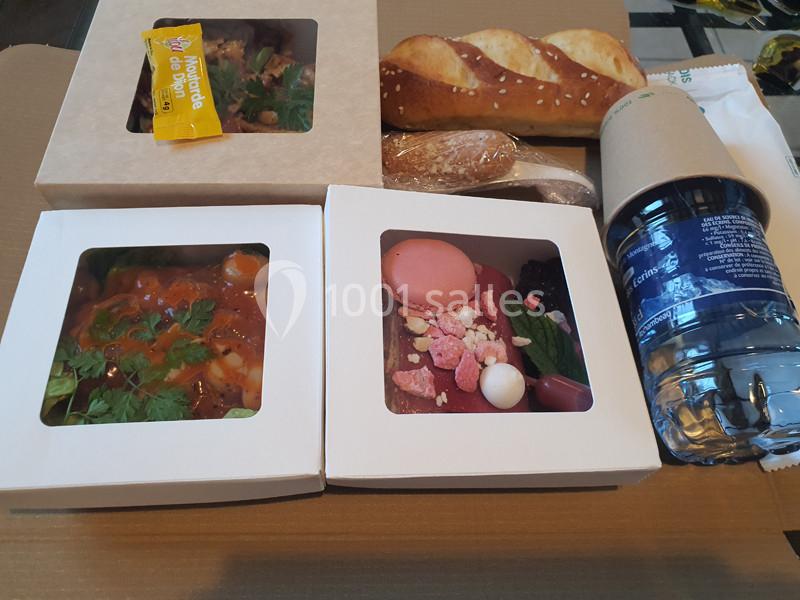 Repas à emporter comprenant plats cuisinés, desserts, baguette, bouteille d'eau et couverts emballés.