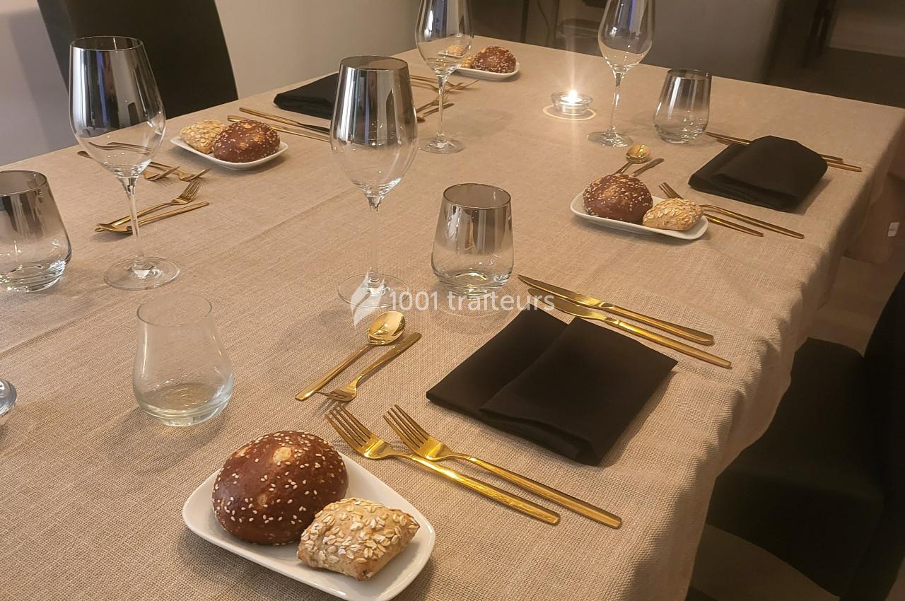 Table dressée avec une nappe beige, couverts dorés, serviettes noires, verres et assiettes contenant des petits pains.