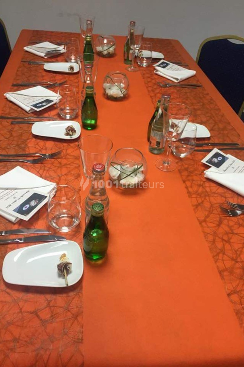 Table dressée avec nappe orange, couverts, verres, bouteilles et assiettes, disposée pour un repas ou une réunion.