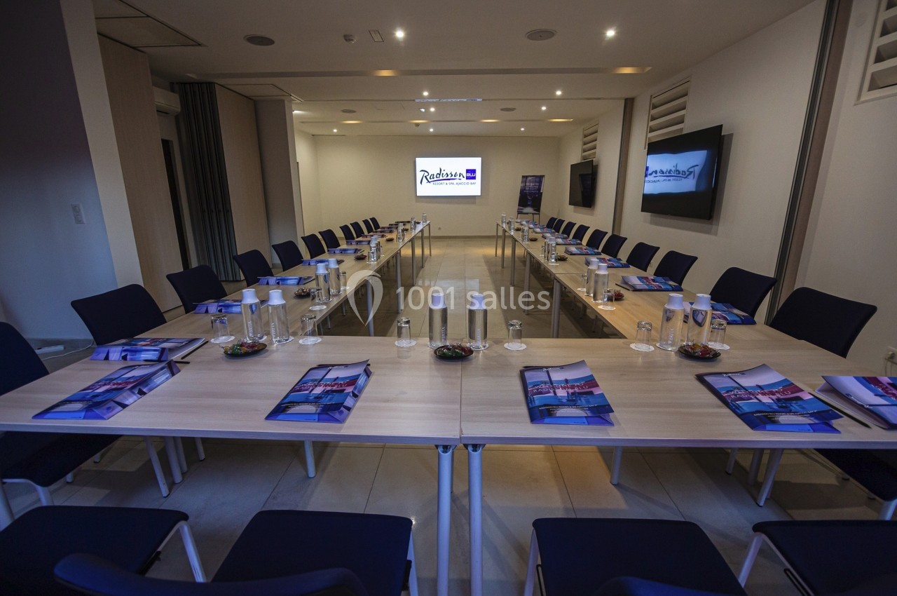 Salle de réunion moderne avec tables disposées en U, chaises, documents, bouteilles d'eau et écran de présentation allumé.