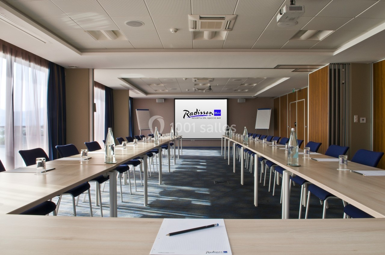 Salle de réunion lumineuse avec tables en U, chaises bleues, bouteilles d'eau et écran de projection au fond.