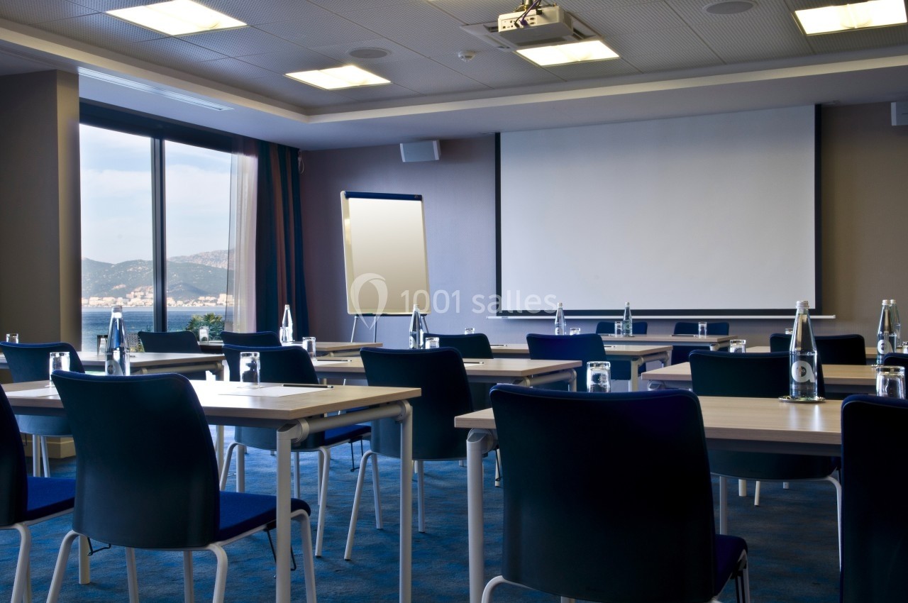 Salle de réunion lumineuse avec tables, chaises, tableau blanc et écran de projection, vue sur l'extérieur.