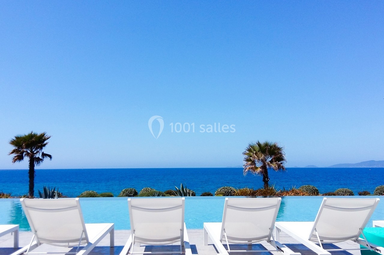 Chaises longues blanches alignées face à une piscine à débordement avec vue sur la mer et deux palmiers.