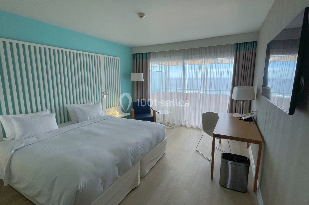 Chambre lumineuse avec lit double, bureau, fauteuil, grande fenêtre et vue sur l'extérieur.