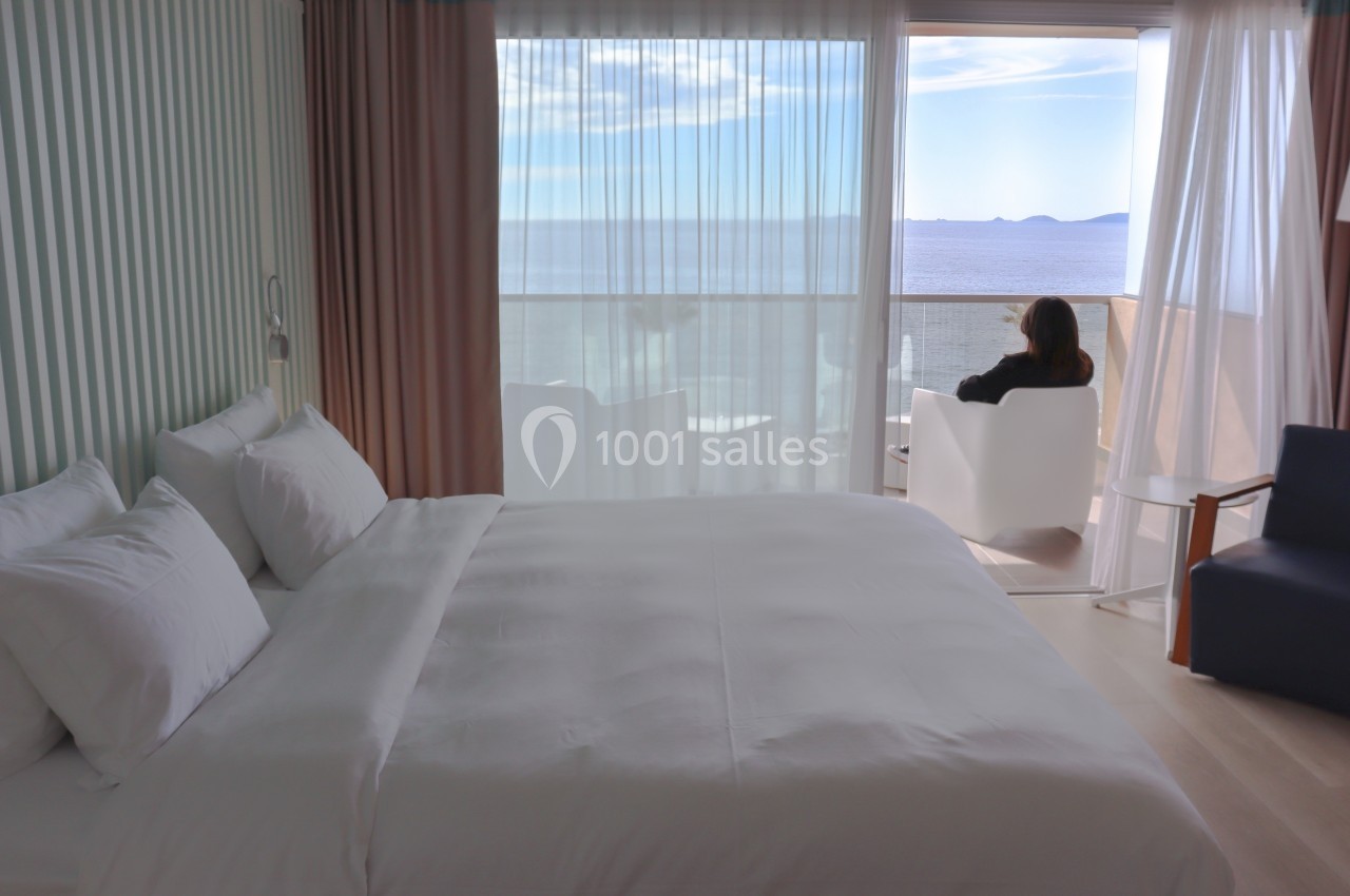 Chambre lumineuse avec lit double, rideaux ouverts sur une baie vitrée offrant une vue sur la mer, personne assise au balcon.