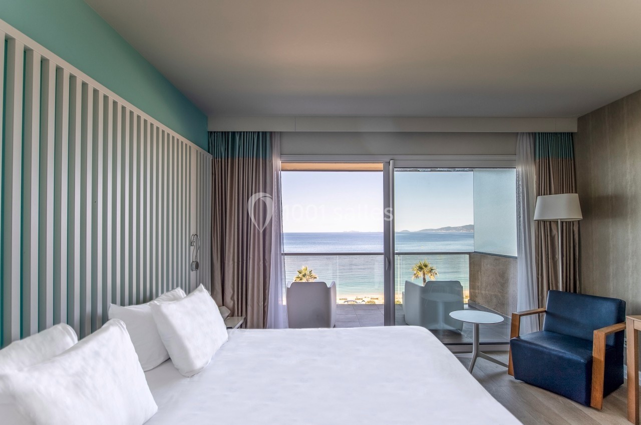 Chambre lumineuse avec lit double, fauteuil, table et balcon offrant une vue sur la mer et un ciel dégagé.