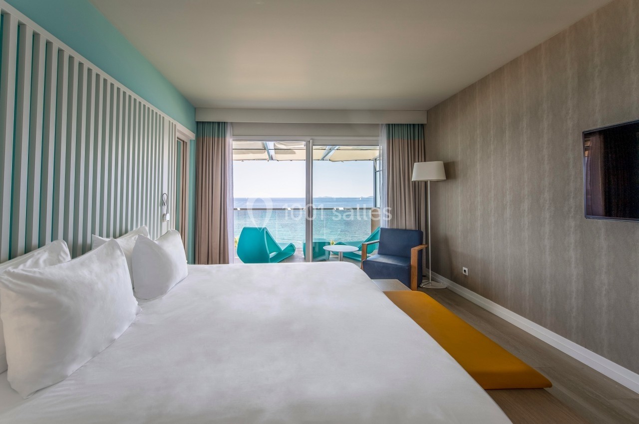 Chambre lumineuse avec lit double, fauteuils, vue sur la mer et balcon équipé de chaises et table.