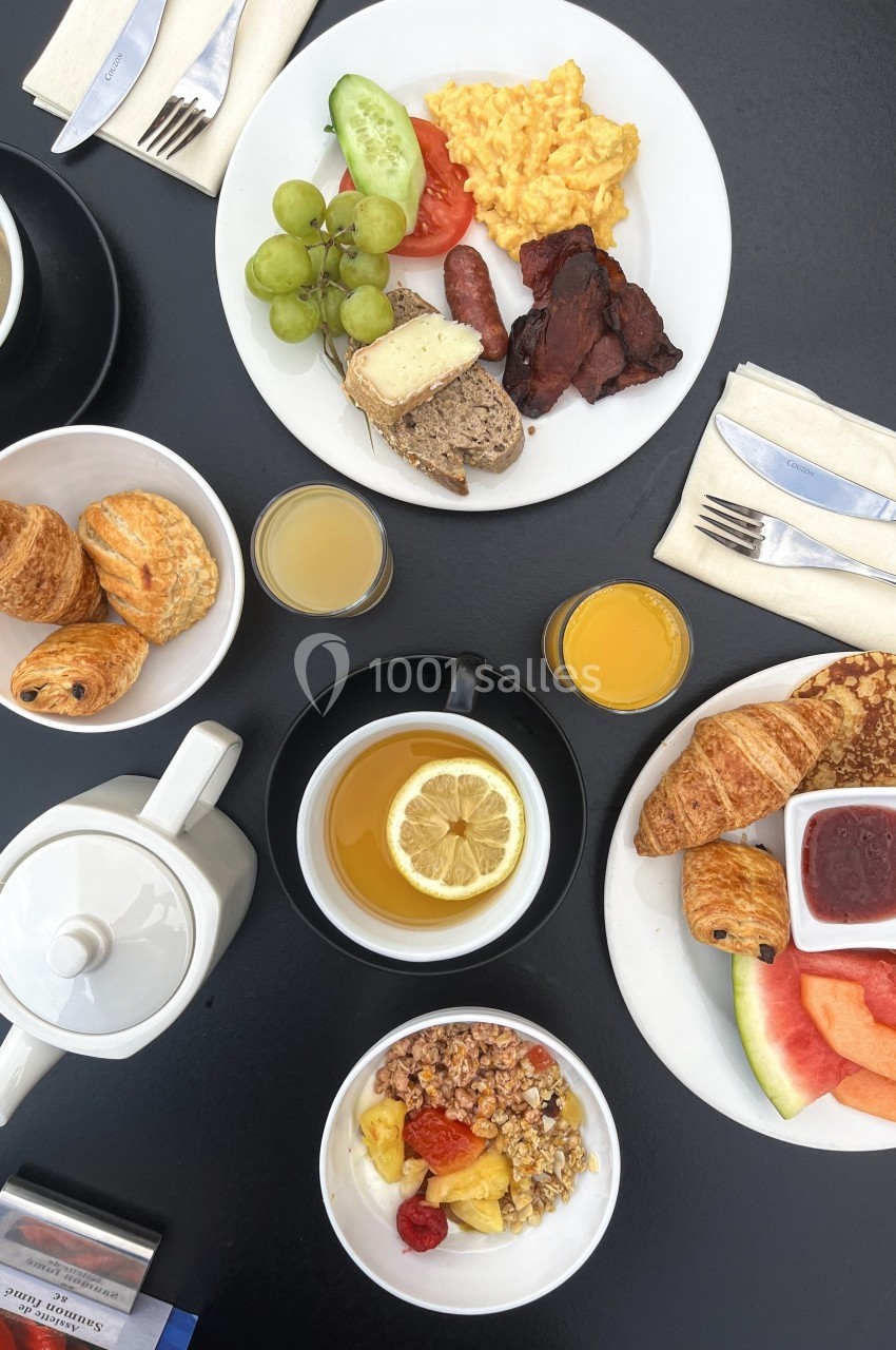 Petit-déjeuner varié avec fruits, viennoiseries, céréales, œufs brouillés, charcuterie et boissons sur une table noire.