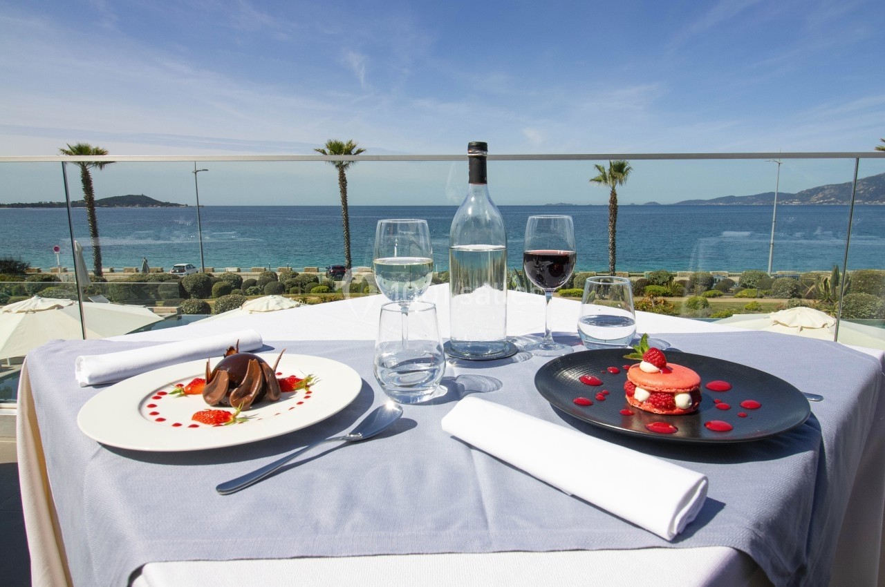 Table dressée avec vue sur la mer, comprenant des desserts élégants, un verre de vin rouge et une bouteille d'eau.