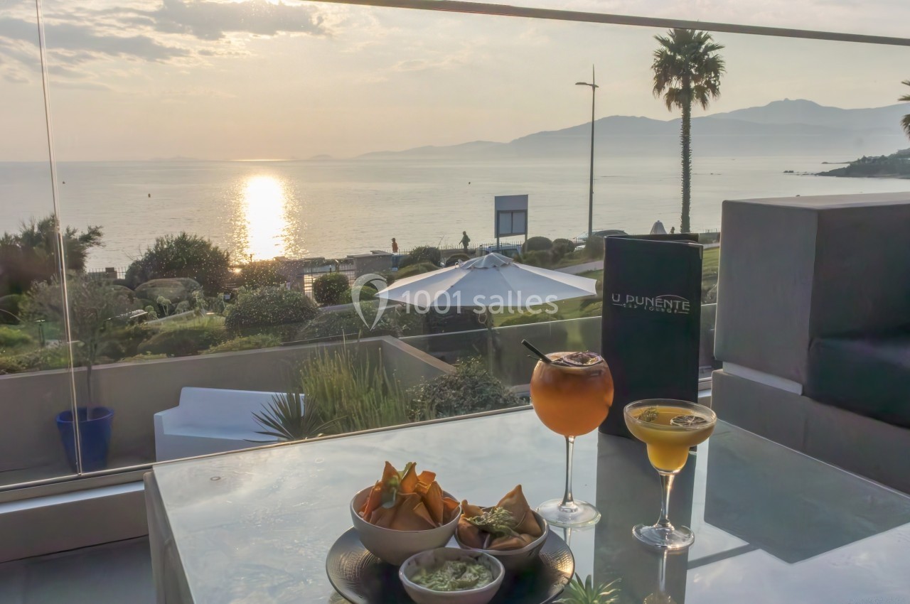 Deux cocktails et des amuse-bouches sur une table en terrasse avec vue sur la mer au coucher du soleil.