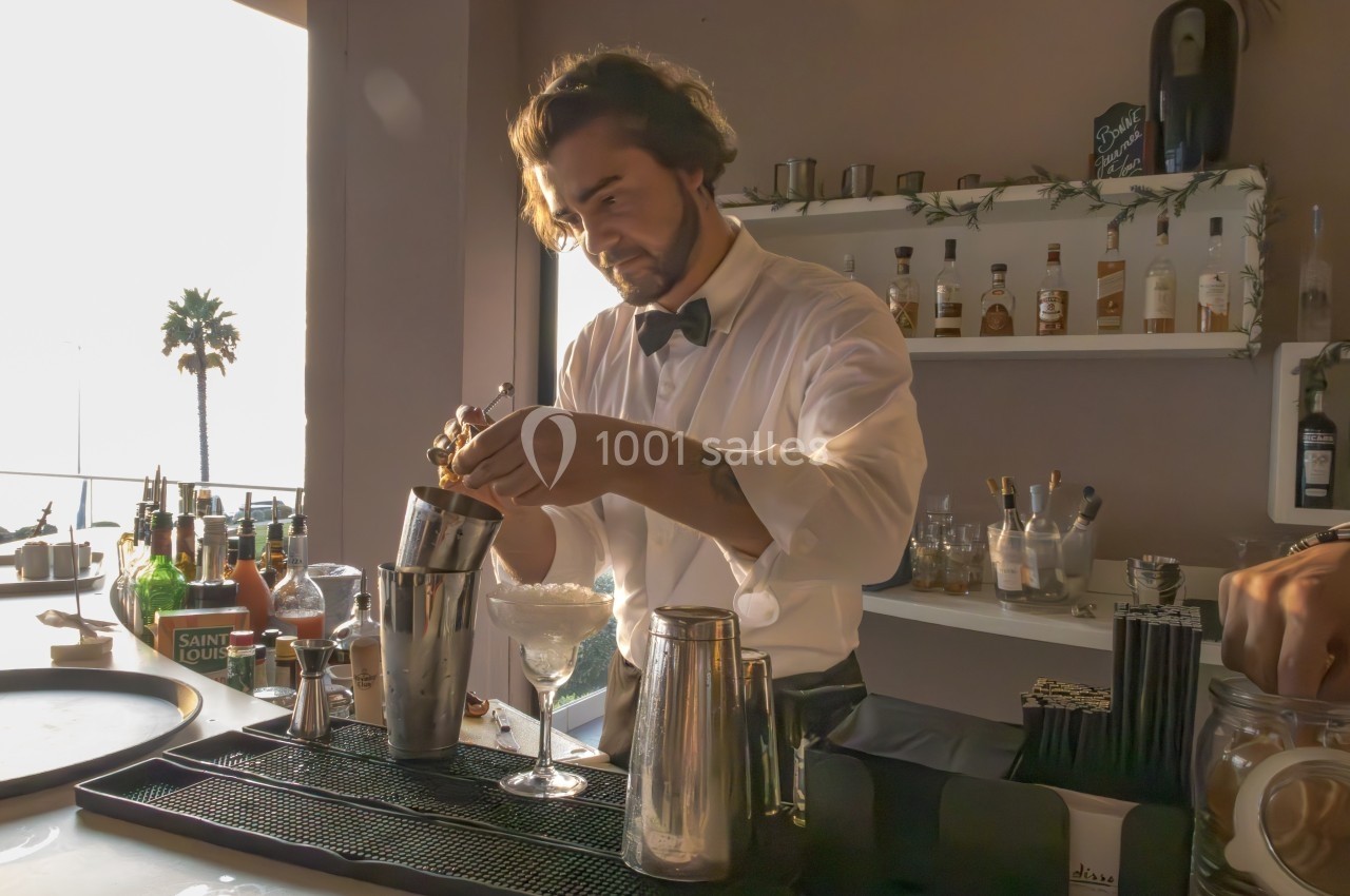 Un barman en chemise blanche prépare un cocktail dans un shaker sur un comptoir de bar ensoleillé.