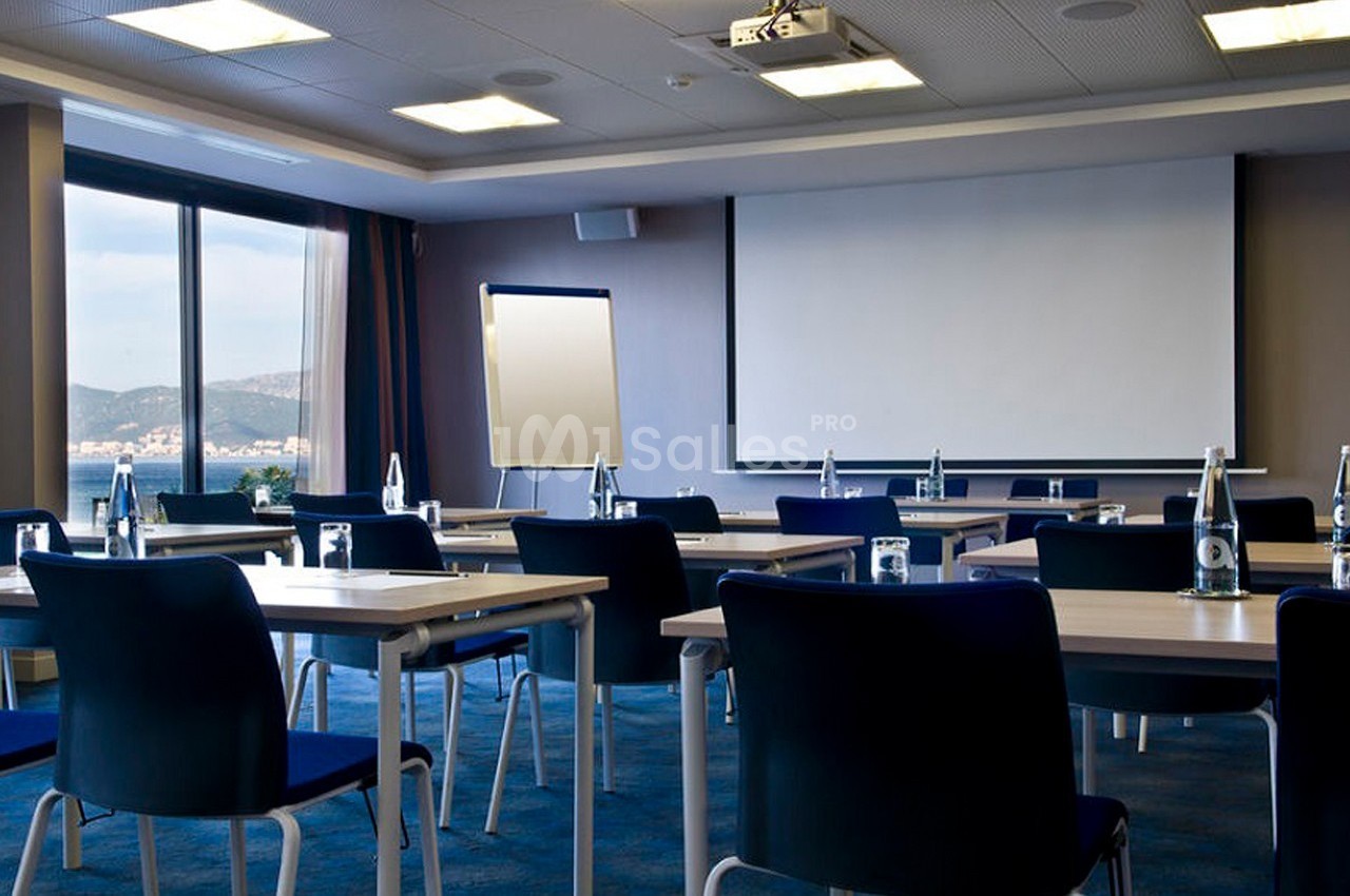 Salle de réunion lumineuse avec tables, chaises, tableau blanc et écran de projection, vue sur l'extérieur.