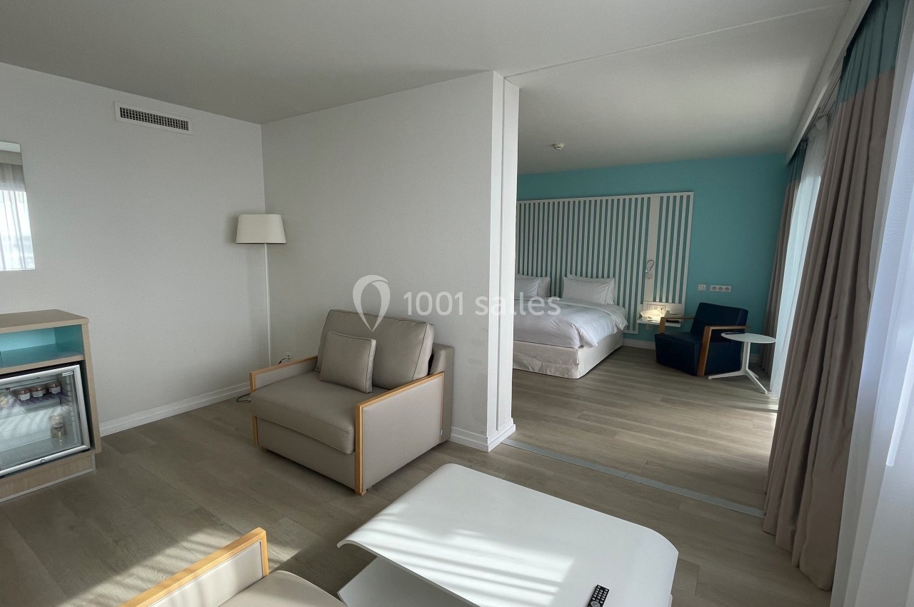 Chambre d'hôtel lumineuse avec coin salon, lit double, mini-réfrigérateur et grandes fenêtres laissant entrer la lumière…