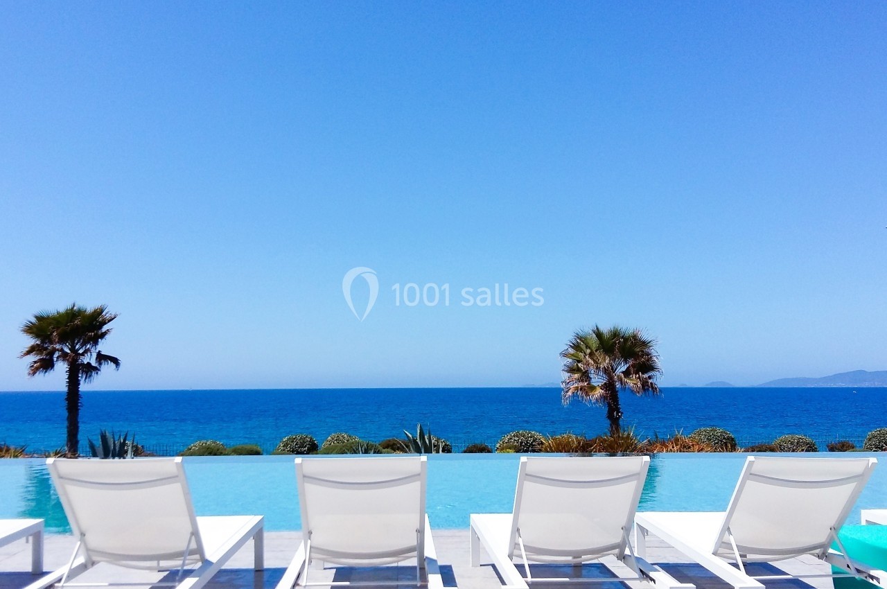 Chaises longues blanches alignées face à une piscine à débordement avec vue sur la mer et deux palmiers.