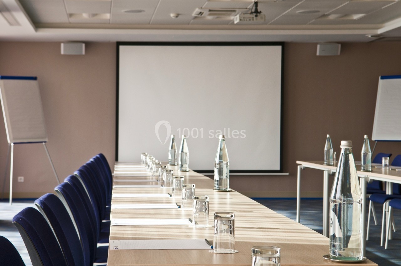 Salle de réunion avec une table longue, des chaises alignées, des bouteilles d'eau, des verres et un écran de projection.