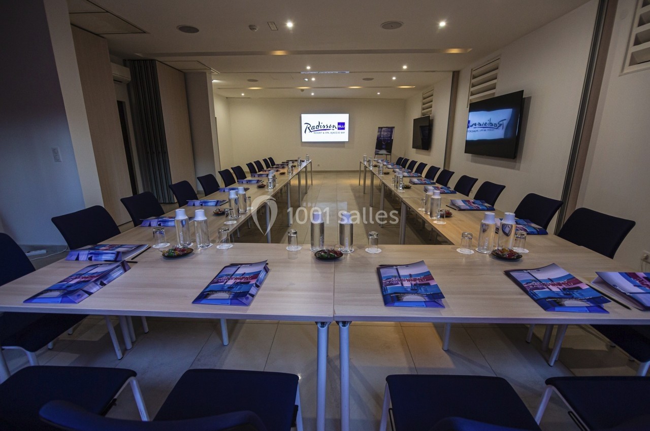 Salle de réunion moderne avec tables disposées en U, chaises, documents, bouteilles d'eau et écran allumé en fond.