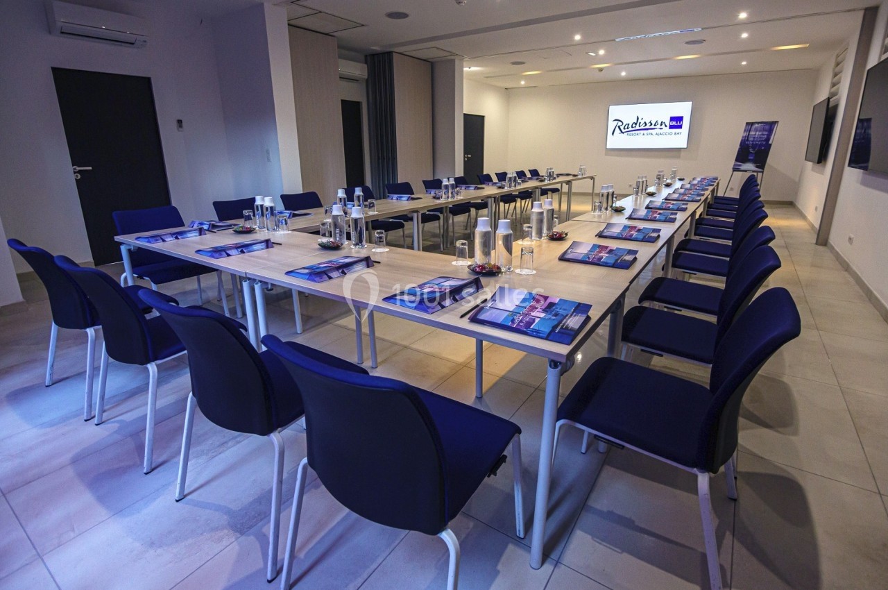 Salle de réunion moderne avec tables disposées en U, chaises bleues et matériel de conférence sur les tables.
