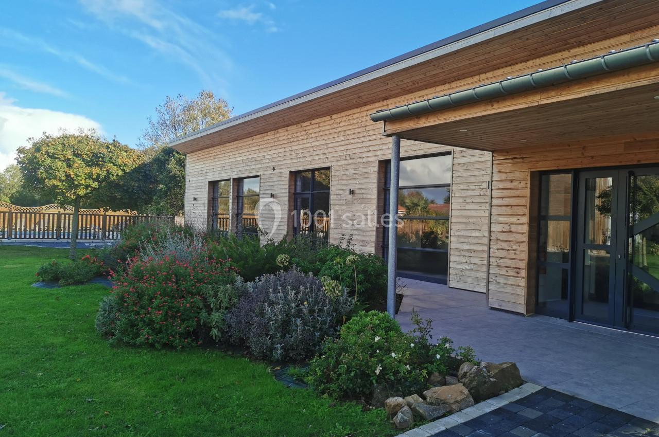 Maison en bois moderne avec terrasse couverte, jardin fleuri et pelouse, sous un ciel bleu dégagé.