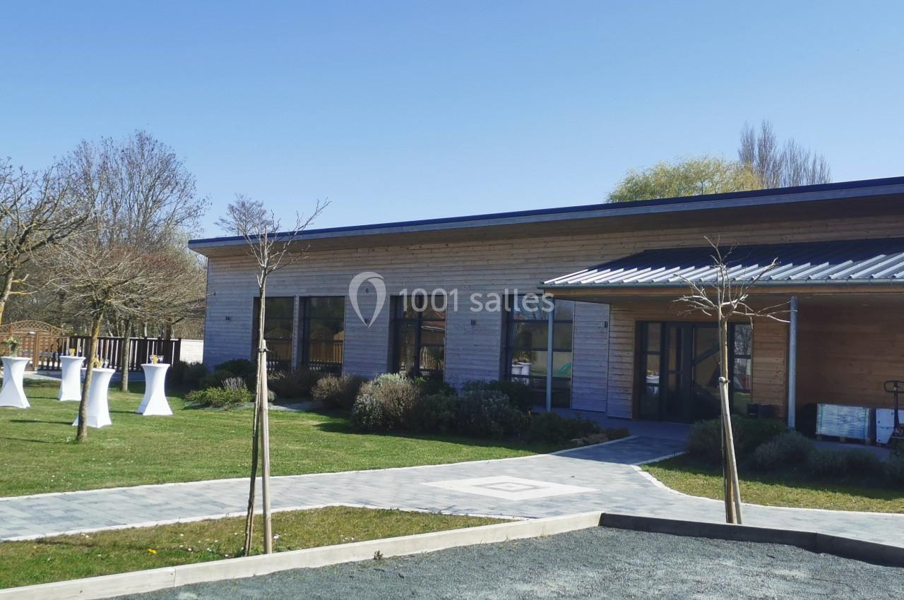 Bâtiment moderne en bois entouré d'un jardin avec des arbres, des tables hautes blanches et un chemin pavé.