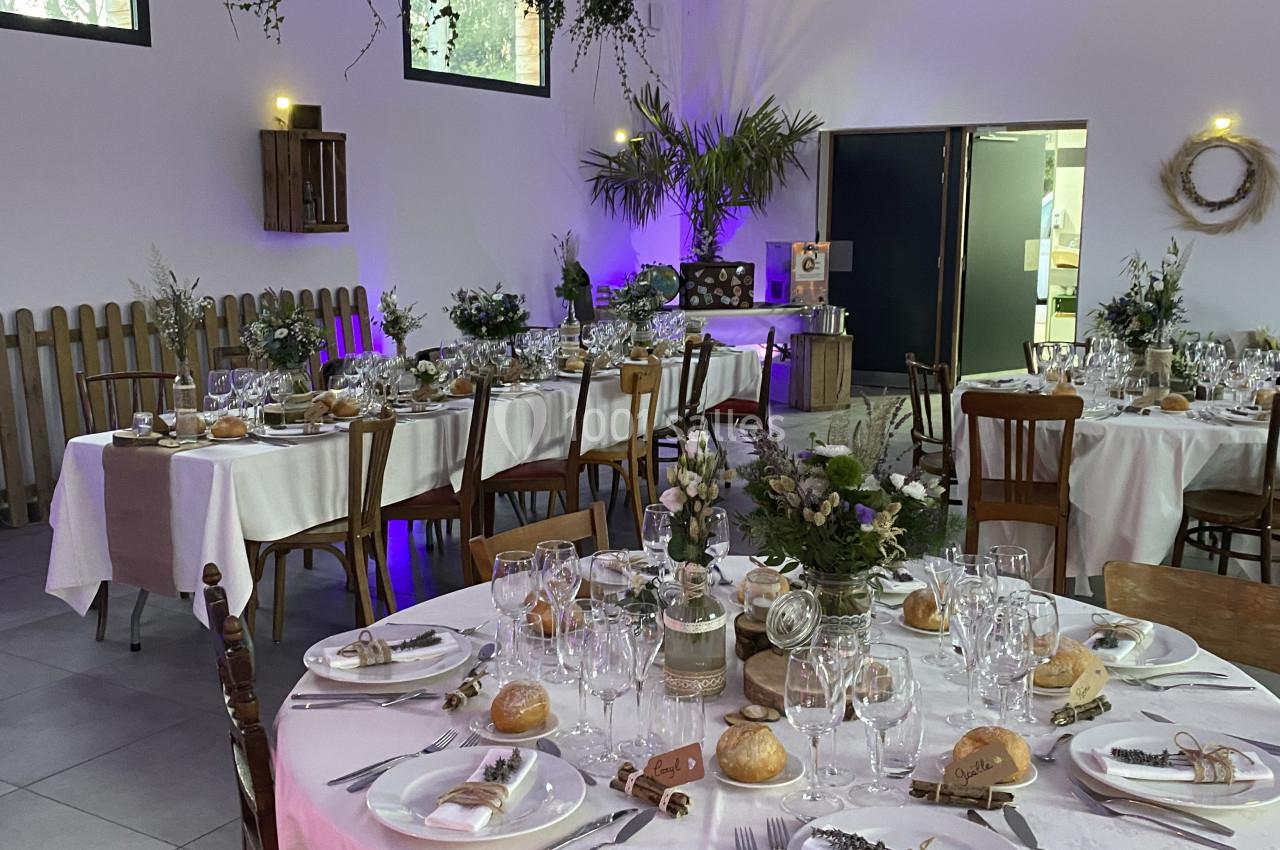 Salle décorée pour un événement avec tables rondes dressées, chaises en bois et végétation suspendue.