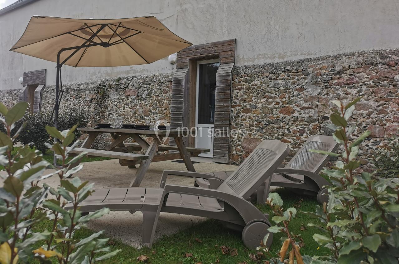 Salon de jardin avec chaises longues, table en bois et parasol beige, devant un mur en pierre et une porte vitrée.