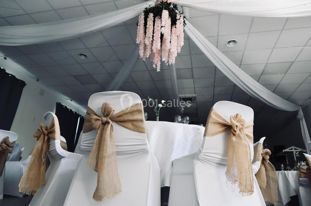 Salle de réception décorée avec des chaises blanches ornées de nœuds en toile de jute et un lustre floral suspendu.