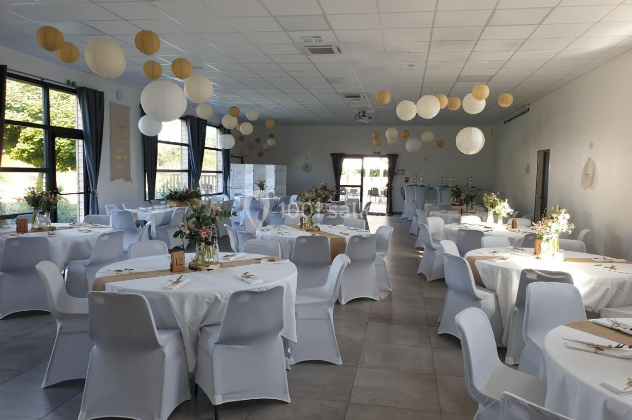 Salle de réception décorée avec des tables rondes, nappes blanches, fleurs et lanternes suspendues.