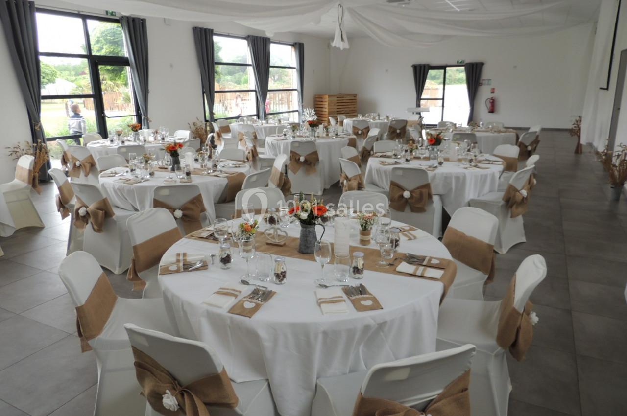 Salle de réception décorée avec des tables rondes, nappes blanches, chaises ornées de nœuds en toile de jute.