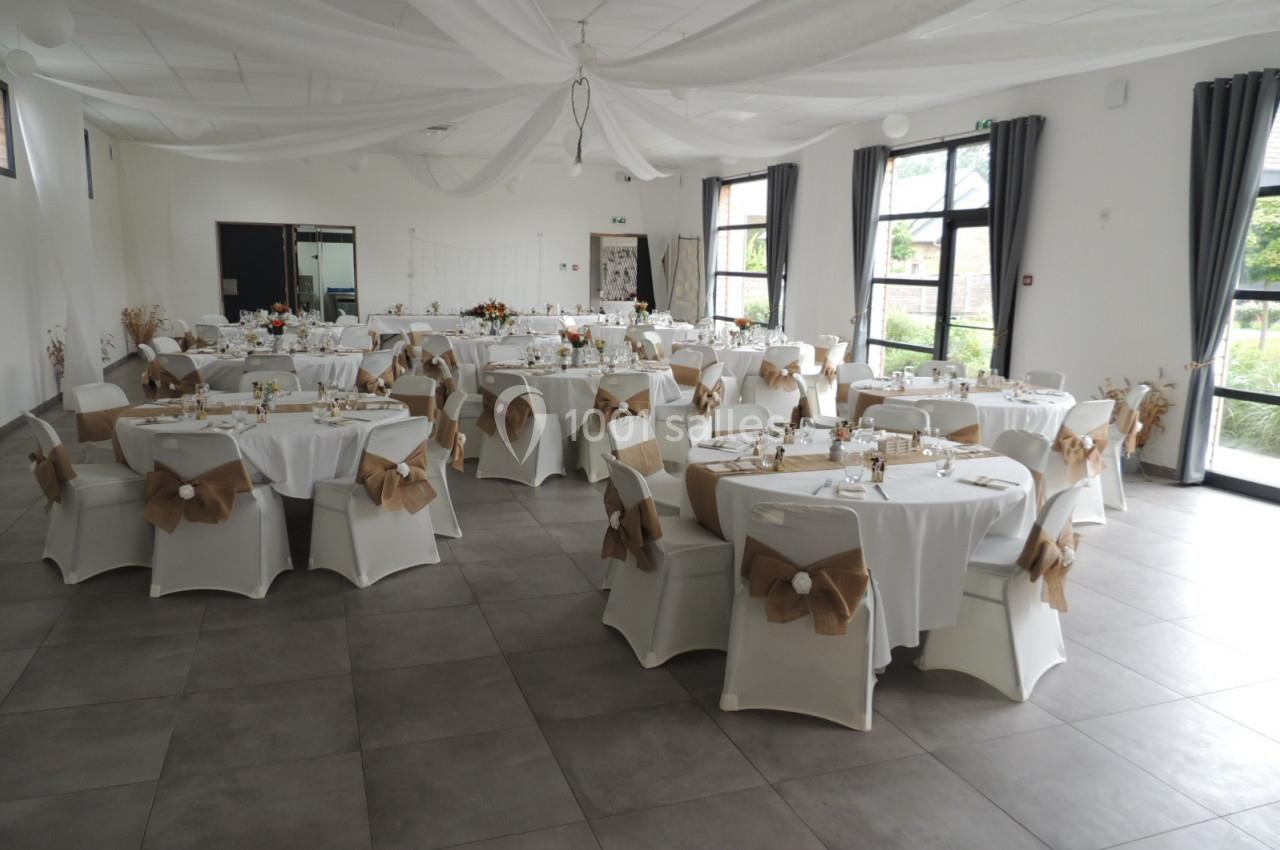 Salle de réception décorée avec des tables rondes, nappes blanches et chaises ornées de nœuds en tissu beige.