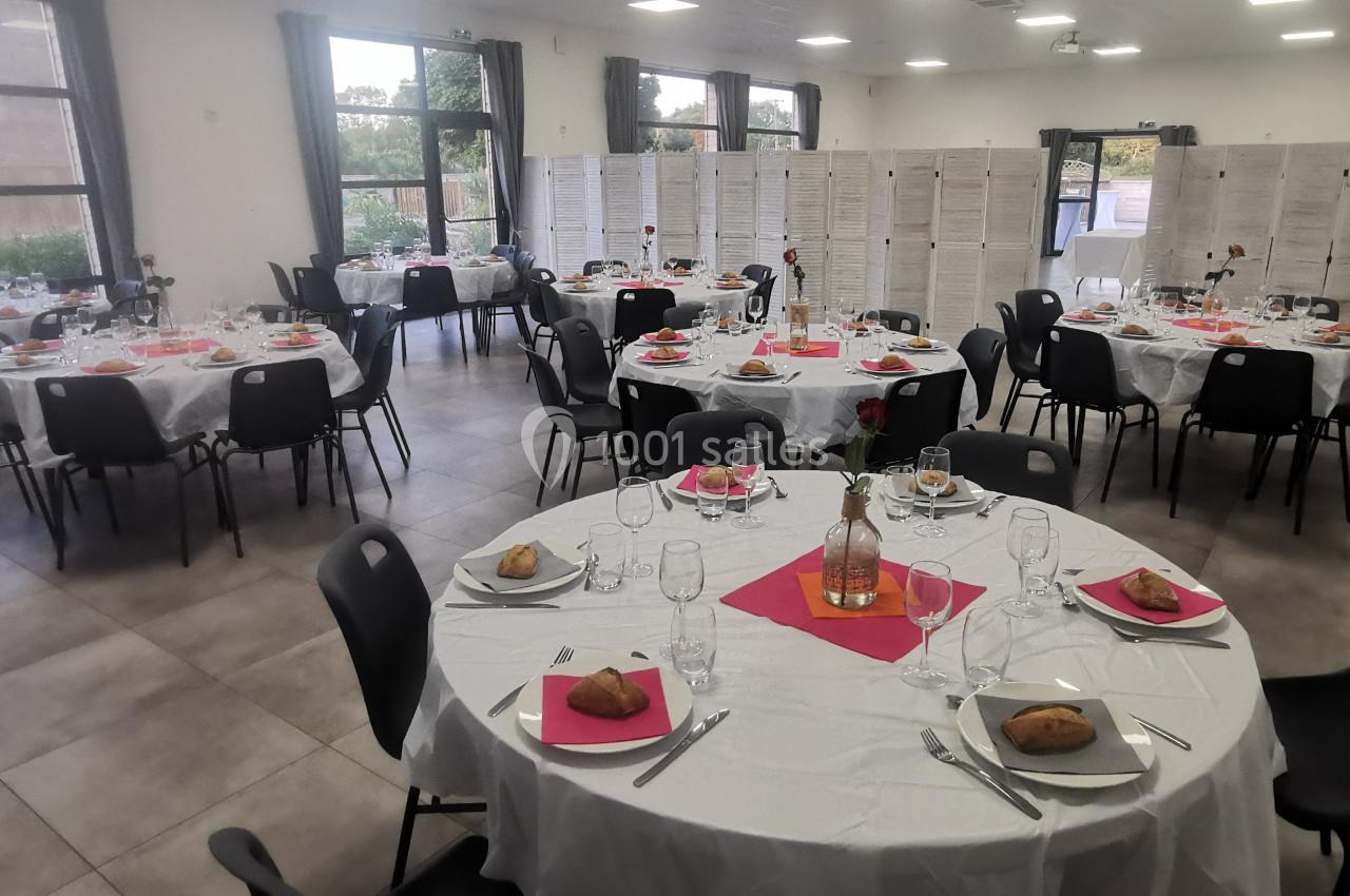 Salle de réception avec tables rondes dressées, nappes blanches, serviettes roses et vaisselle, prête pour un événement.