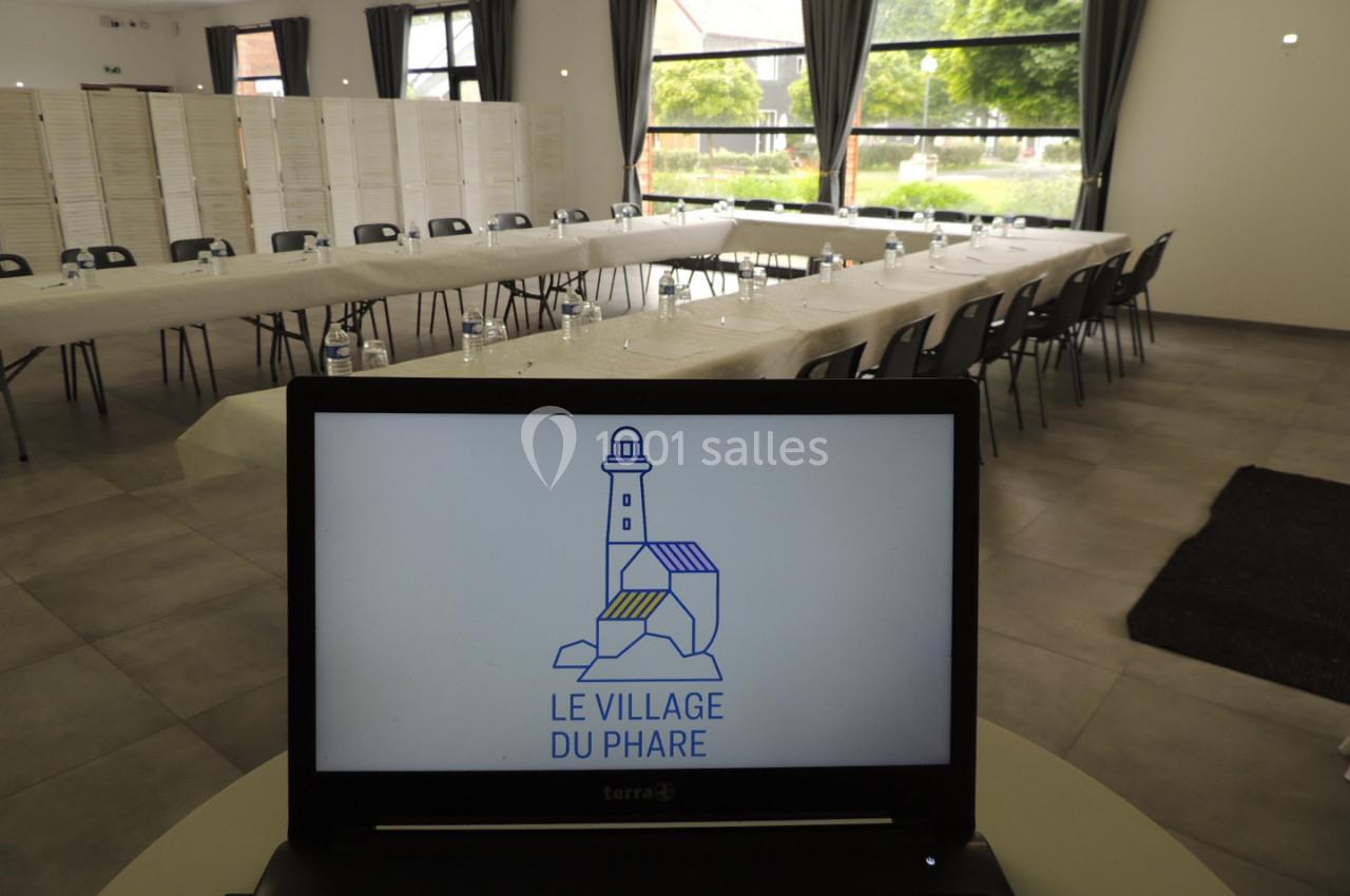 Salle de réunion lumineuse avec tables en U, chaises noires et un ordinateur affichant un logo ’Le Village du Phare’.