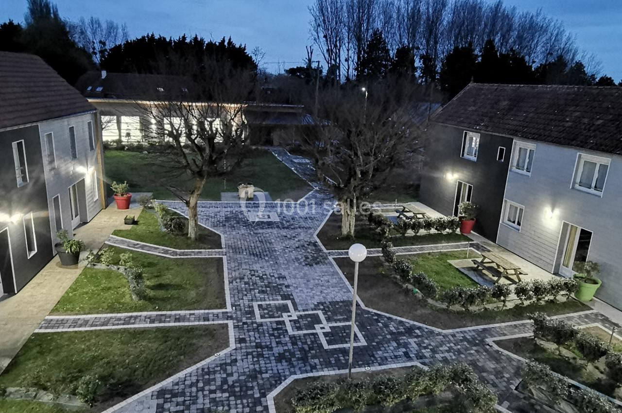 Cour pavée entourée de bâtiments modernes avec espaces verts, arbres et éclairage extérieur au crépuscule.