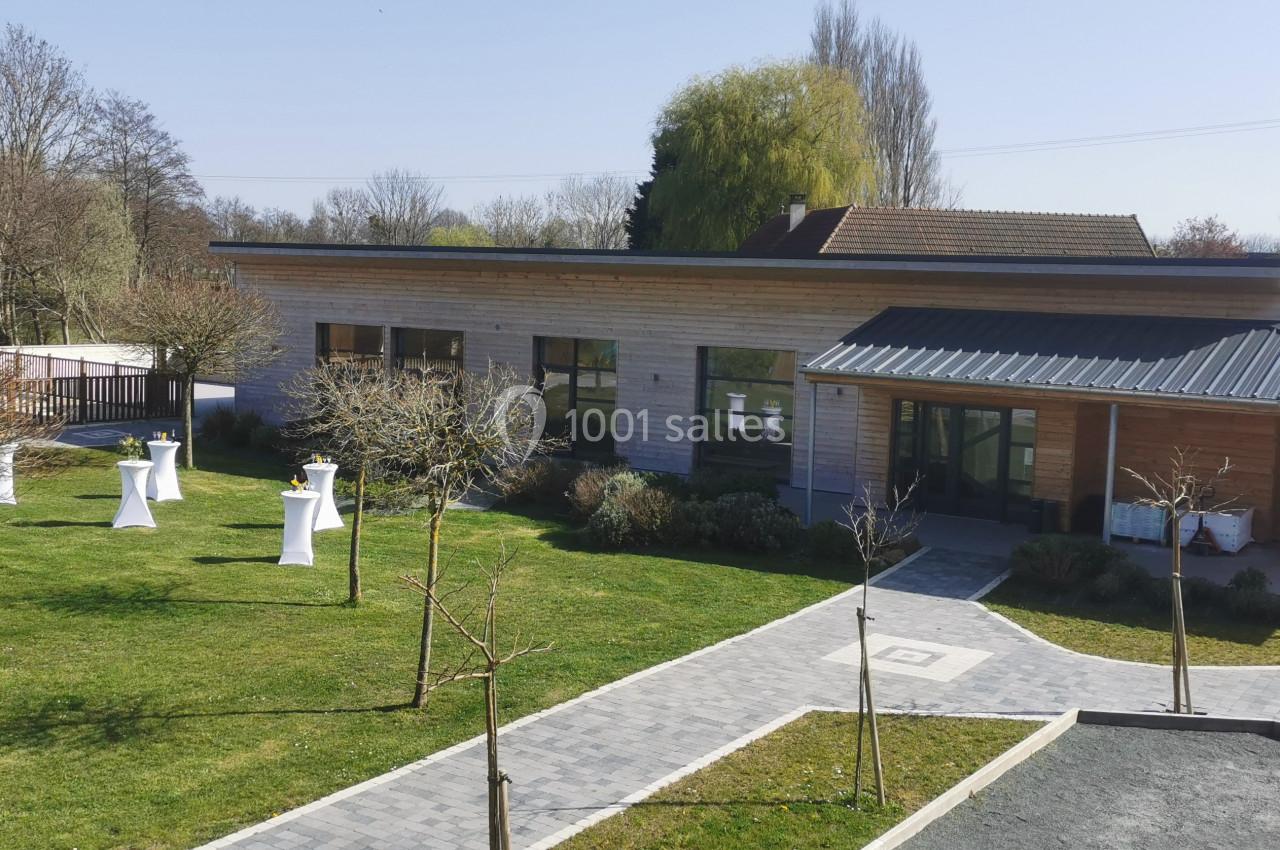 Bâtiment moderne en bois entouré d'un jardin avec des arbres et des tables hautes blanches en extérieur.
