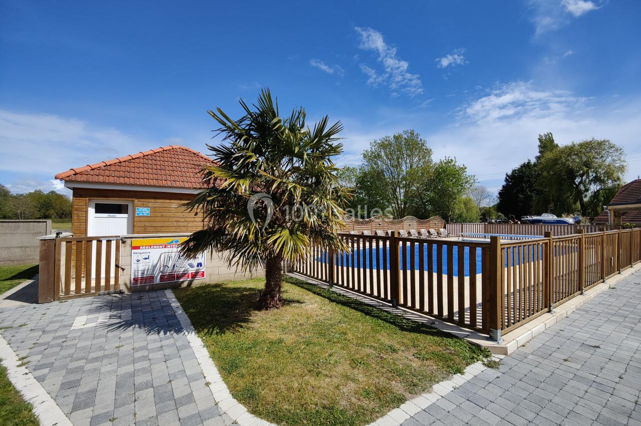 Piscine extérieure clôturée avec un bâtiment en bois, un palmier et un chemin pavé sous un ciel bleu.