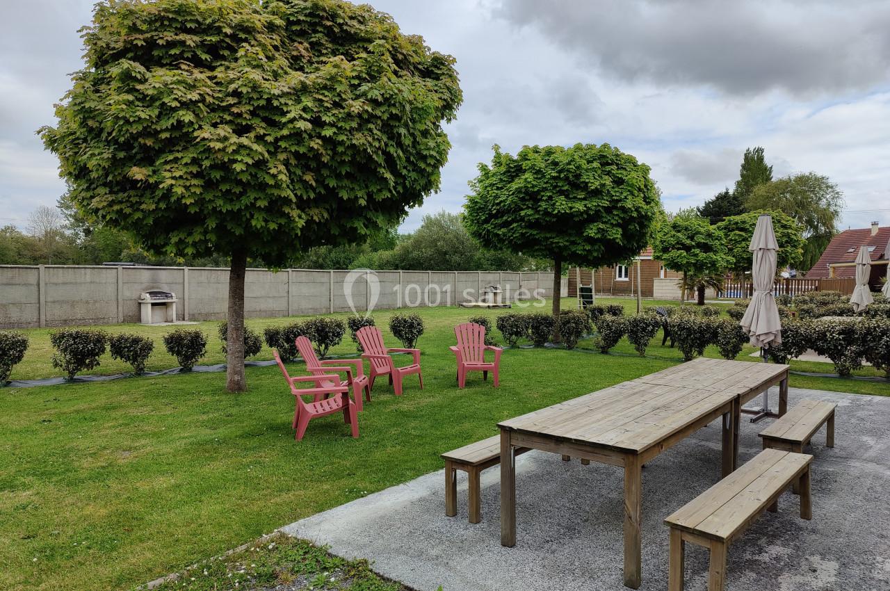 Table en bois avec bancs, chaises roses et parasol dans un jardin arboré, entouré de haies et clôtures grises.