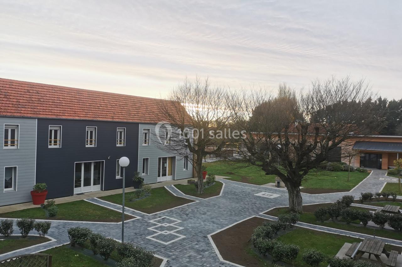 Vue d'une cour pavée avec bâtiments modernes, arbres taillés et bancs, sous un ciel partiellement nuageux.