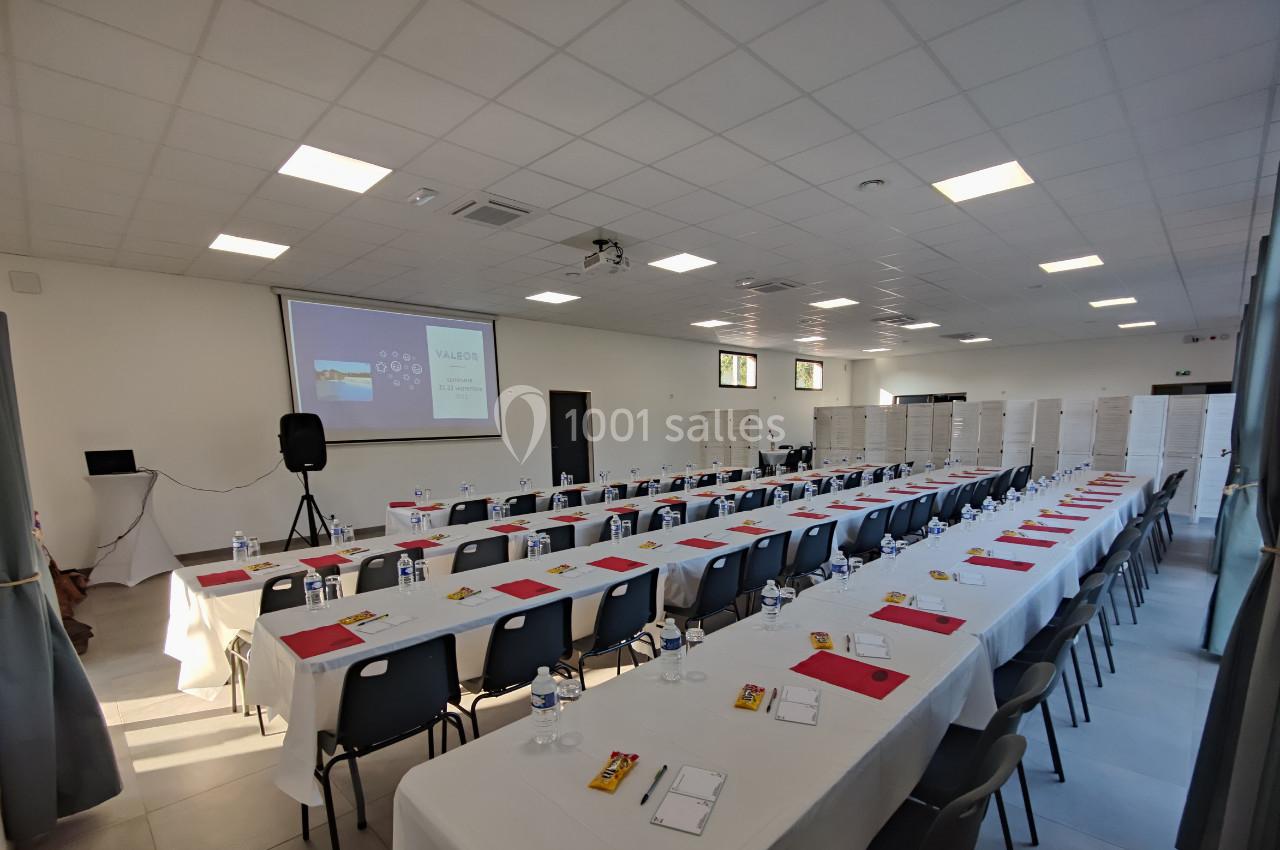 Salle de conférence avec des rangées de tables et chaises, écran de projection allumé et documents disposés sur les tables.