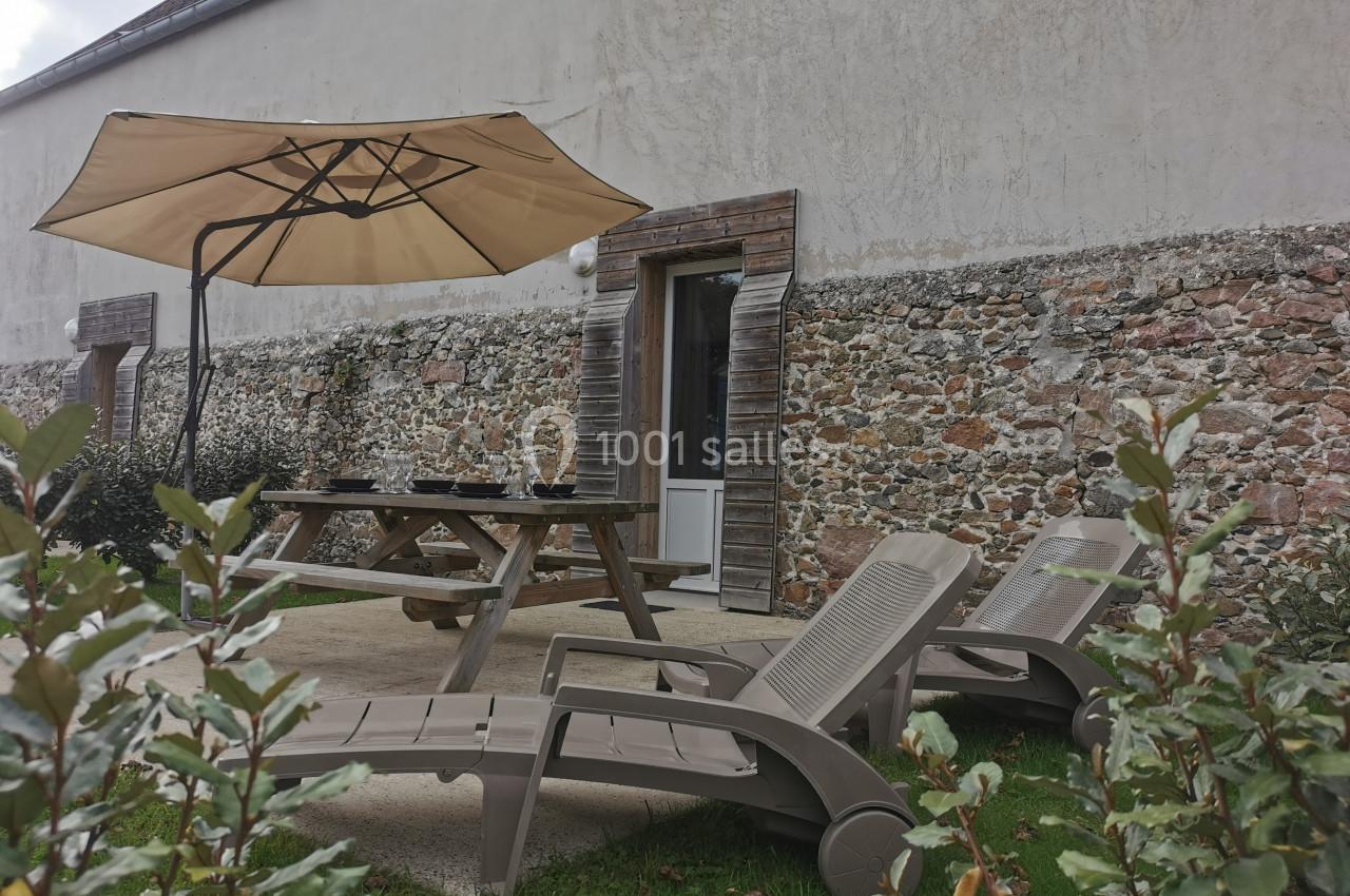 Table de pique-nique et chaises longues disposées sur une terrasse en béton devant un mur en pierre, avec un parasol beige.