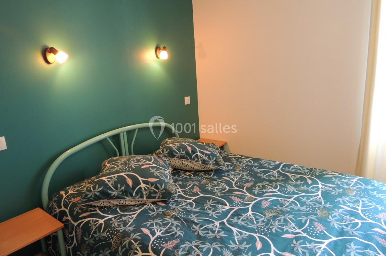 Lit double avec une tête de lit en métal vert, draps à motifs floraux, éclairé par deux appliques murales.