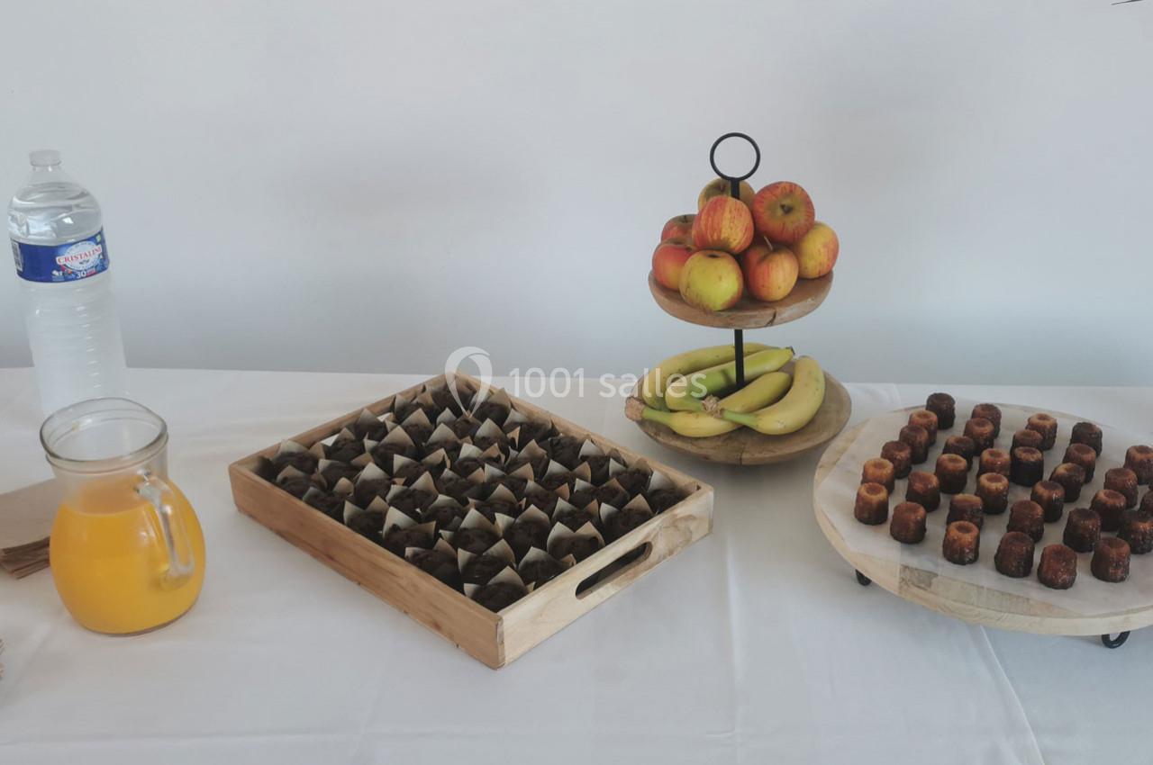Table avec jus d'orange, bouteille d'eau, fruits, cannelés et petits gâteaux disposés sur des plateaux.