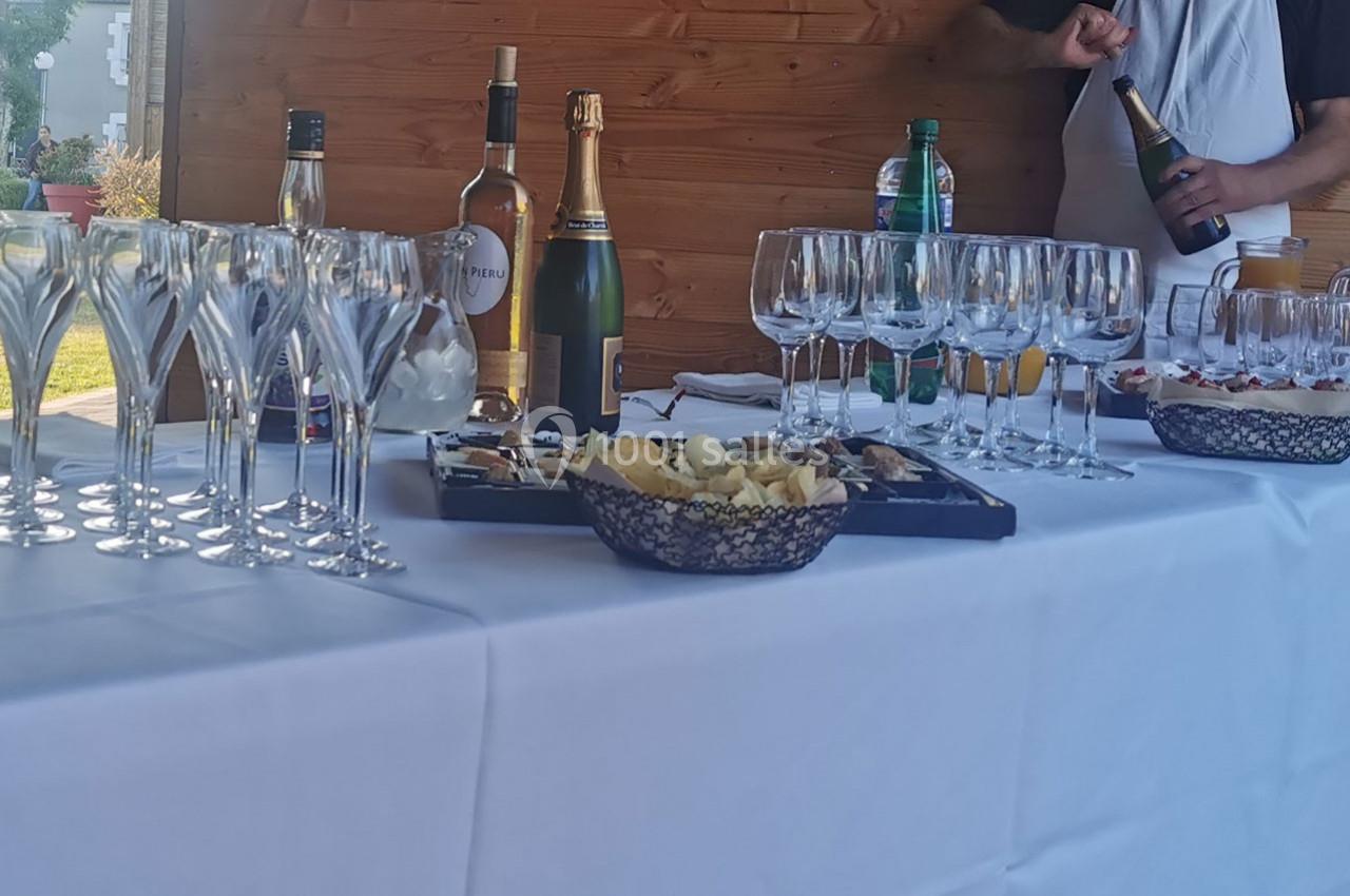Table dressée avec verres, bouteilles de boissons, plateaux de fromages et apéritifs, dans un cadre extérieur.